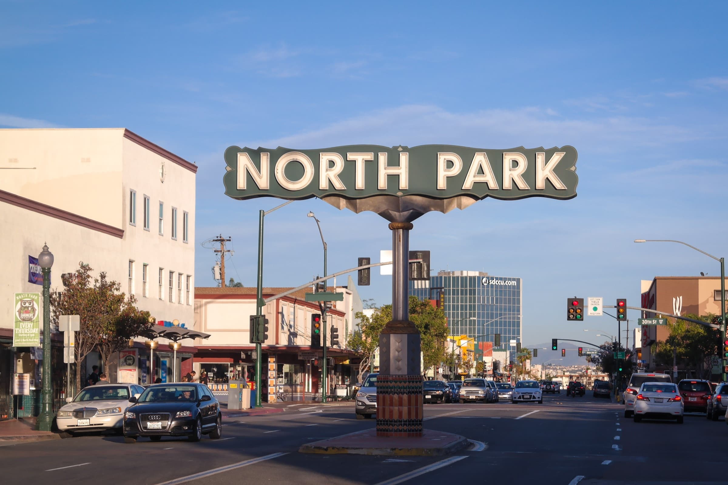 The North Park sign on its tiled column in the median of University Avenue at 30th Street, with two-story storefronts and traffic in late-afternoon light